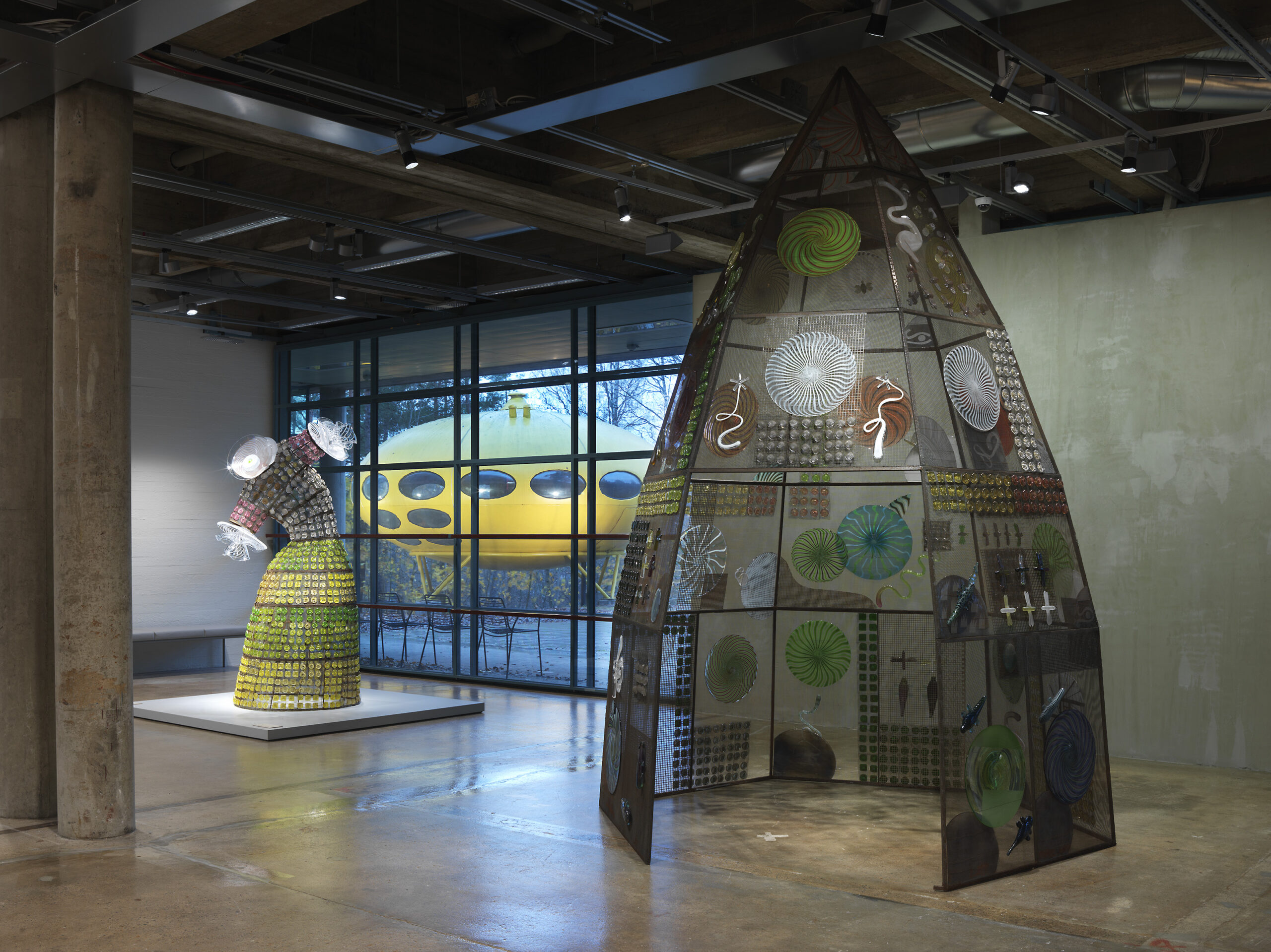 Two large works in a spacious exhibition space. The works, resembling a dress and a gazebo, consist of glass elements attached to a metal frame.