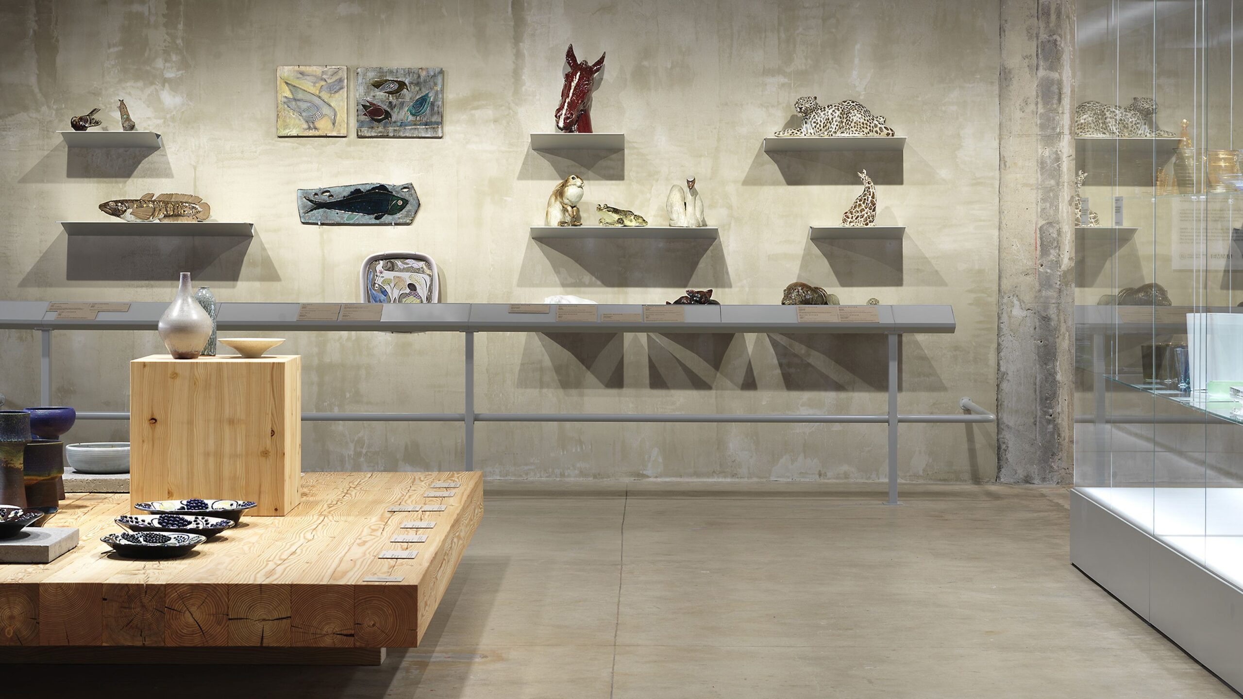 Avara betoninen näyttelytila, jossa on keramiikkateoksia puisella tasolla ja seinälle nostettuna, sekä lasinen vitriini.